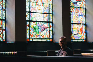 man sitting in empty church pew. Church is dark but light is coming in through stain glass window behind him