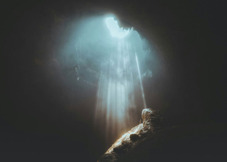 photo of a dark cloudy sky with a burst of light breaking through the clouds onto a rocky mountaintop where a man stands
