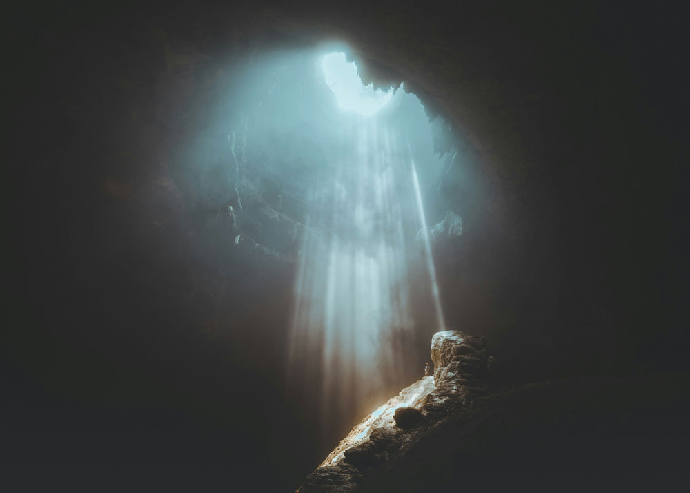 photo of a dark cloudy sky with a burst of light breaking through the clouds onto a rocky mountaintop where a man stands