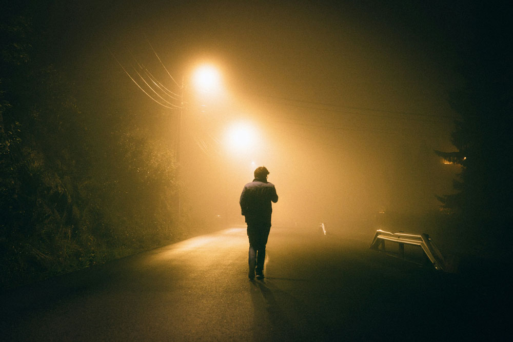 shadow of man walks alone on a dark street that is dimly illuminated by a couple streetlights